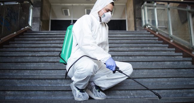 Person in white chemical protection suit disinfecting public corridors and steps to stop spreading highly contagious corona virus.